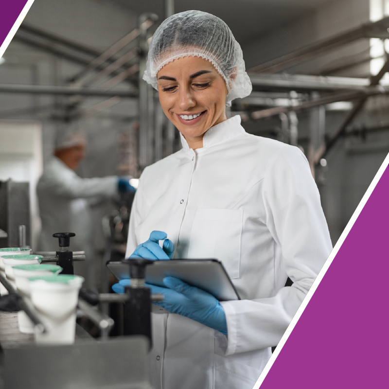 A smiling woman in a white lab coat, hair net, and blue gloves uses a tablet in a clean pharmaceutical or food processing facility. The background shows a blurred industrial setting with another worker and equipment, framed by purple graphic corner accents.
